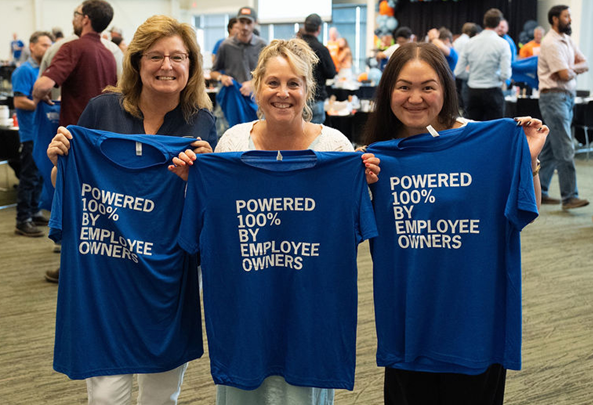 Three women holding blue t-shirts reading Powered 100% by Employee Owners