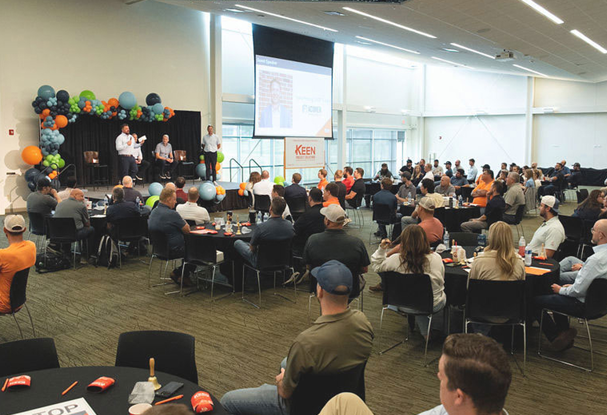 Large group of people sitting around many round tables, listening to a presentation given on stage by a few individuals