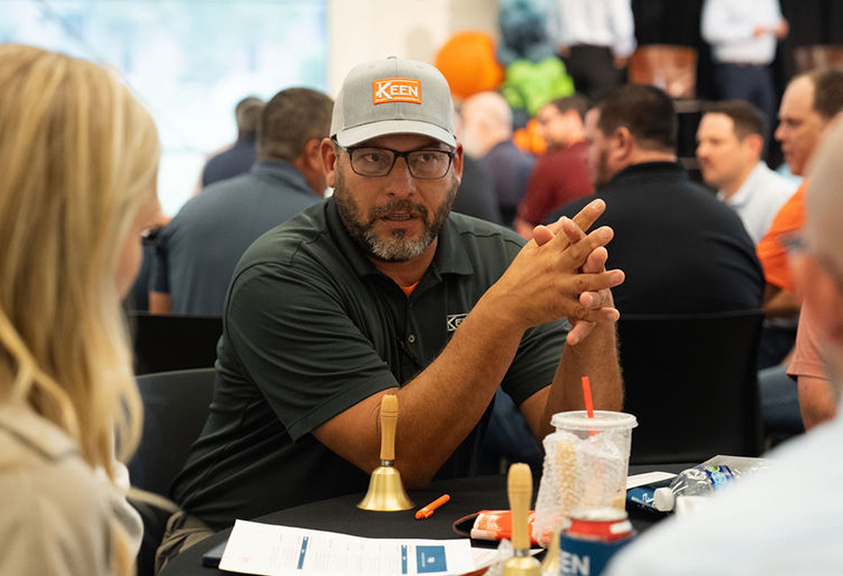 Man wearing a Keen hat, folding his hands on a table, speaking with a few other people