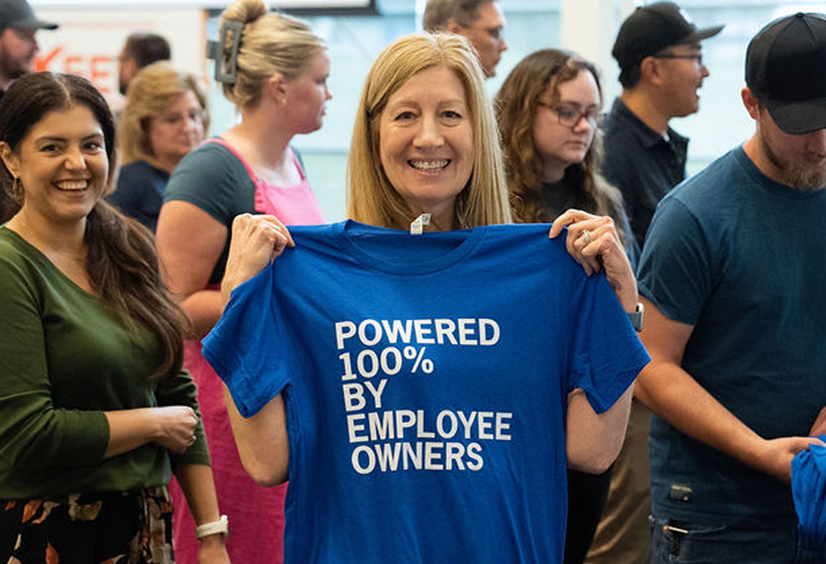 Woman smiling and holding a blue t-shirt reading Powered 100% by Employee Owners, with some people in the background